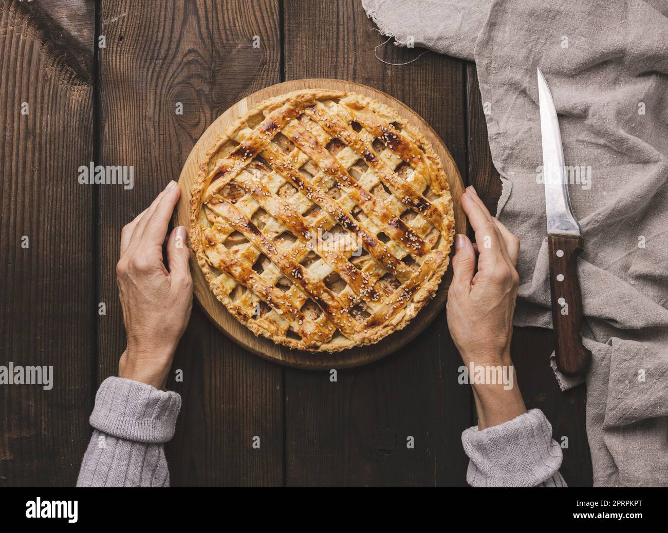 Two female hands hold a round baked pie with apple filling on a wooden board, brown table. Stock Photo