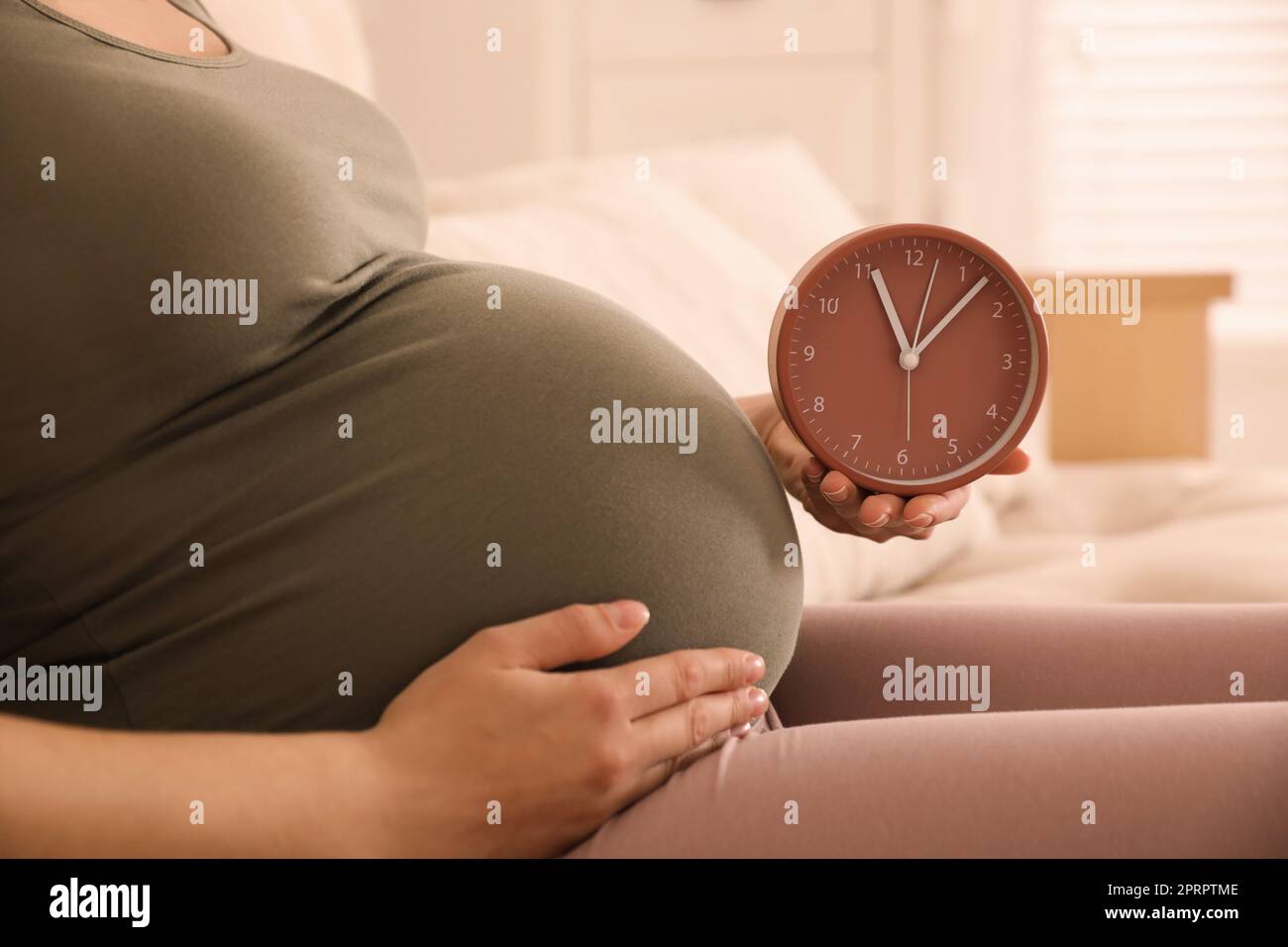 Young pregnant woman holding clock near her belly at home, closeup. Time to give birth Stock Photo