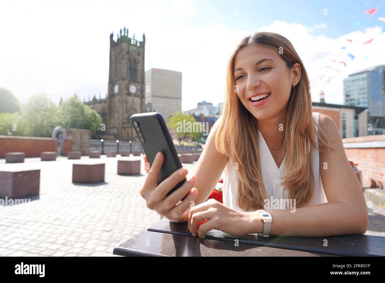 Smiling girl with mobile phone in her hands sits at the table outdoors with the cityscape on the background. Beautiful young woman is watching videos and photos on her smartphone at the cafe table. Stock Photo