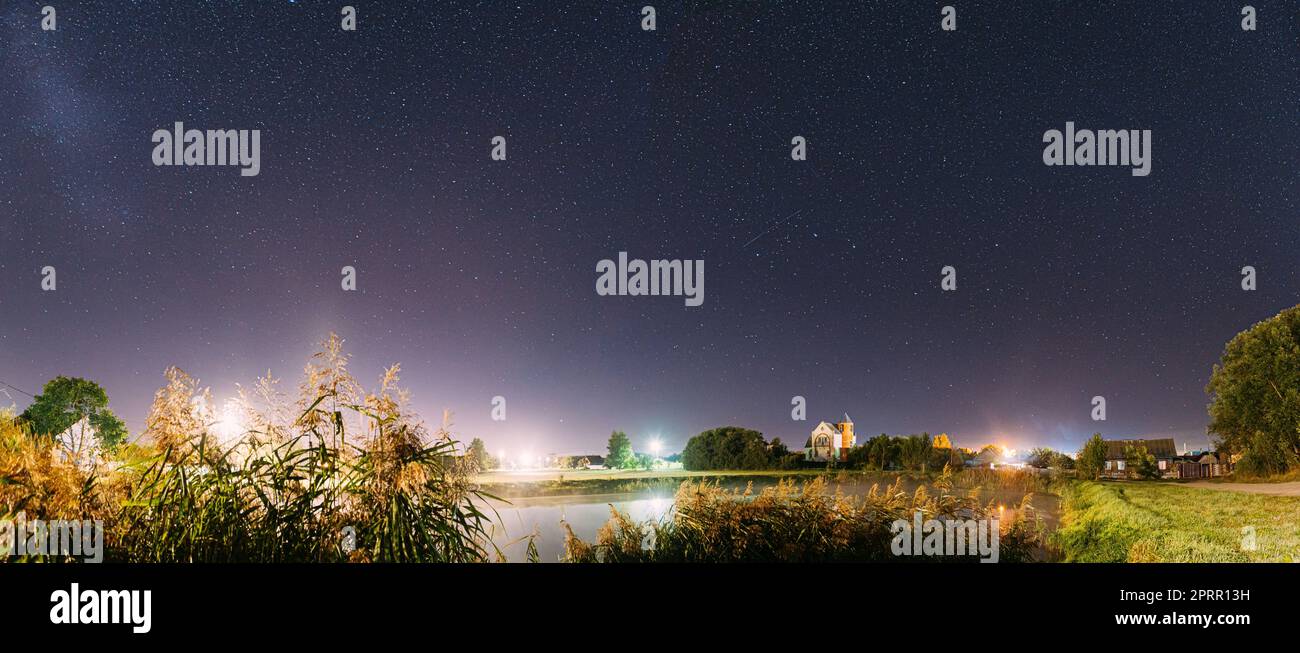 Panorama Of Evaporation Over River Lake Near Houses In Village. Night Starry Sky Above Lake River With Bright Stars And Meteoric Track Trail. Glowing Stars Above Summer Nature Stock Photo