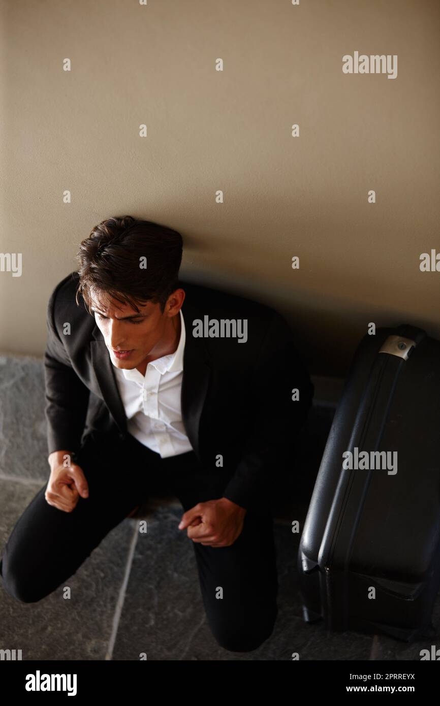 He desperately needs a holiday. an exhausted young businessman sitting on the floor beside his luggage. Stock Photo