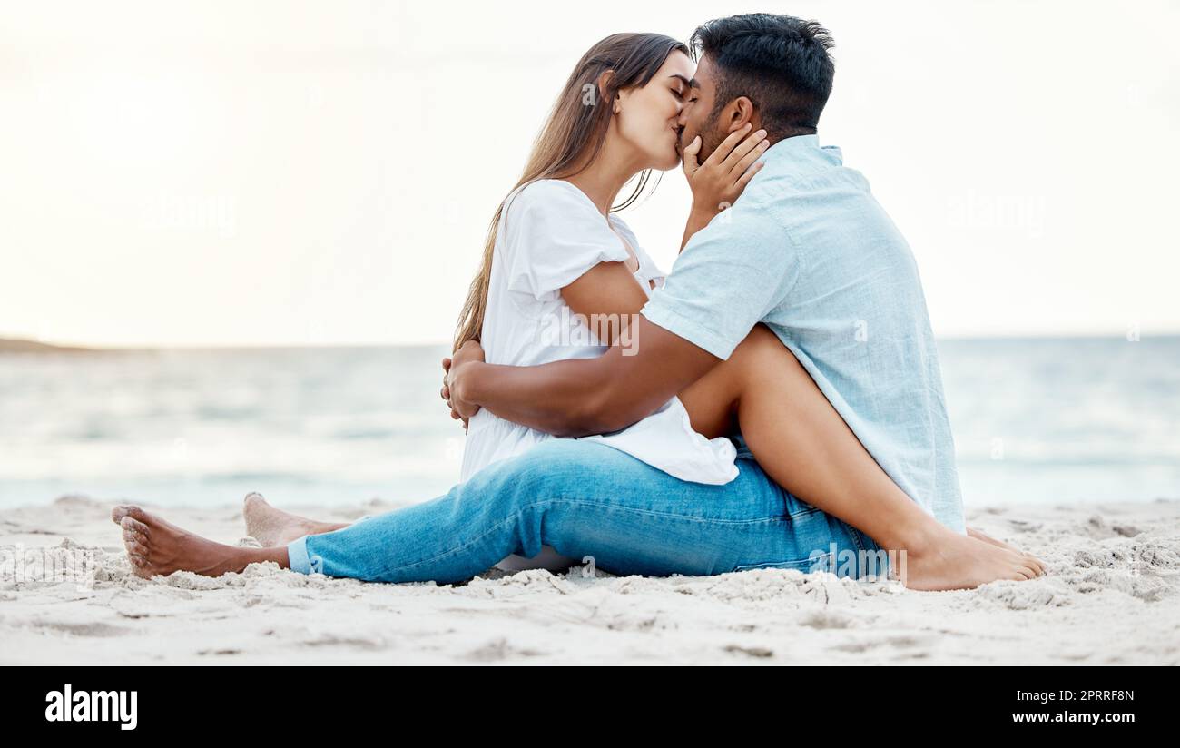 Kiss, beach and love of couple on a date for anniversary, valentines day or romance summer holiday with clear sky, ocean waves and sand. Romantic, intimate and marriage honeymoon or engagement people Stock Photo