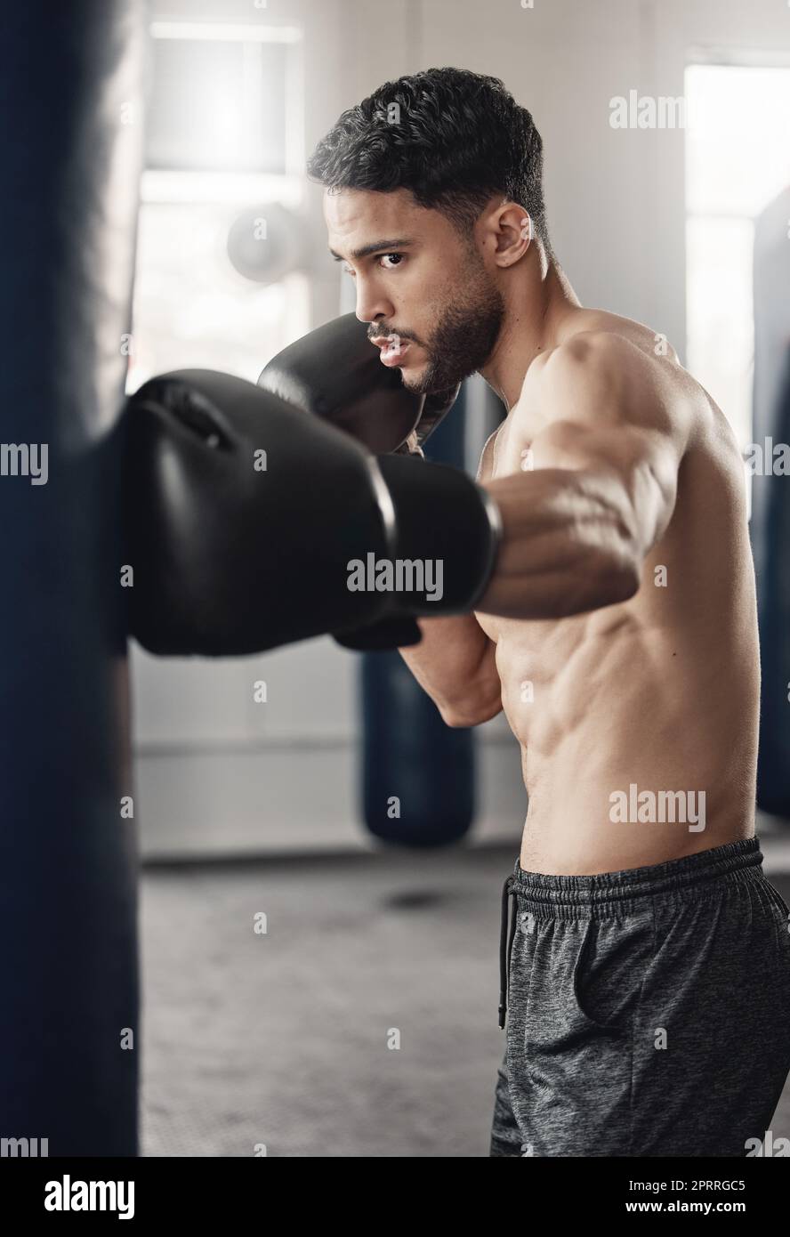 Punching bag, boxing and boxer man in workout training or exercise in a gym. Strong, powerful and serious athlete or personal trainer with fitness gear for muscle strength, wellness or health goals Stock Photo