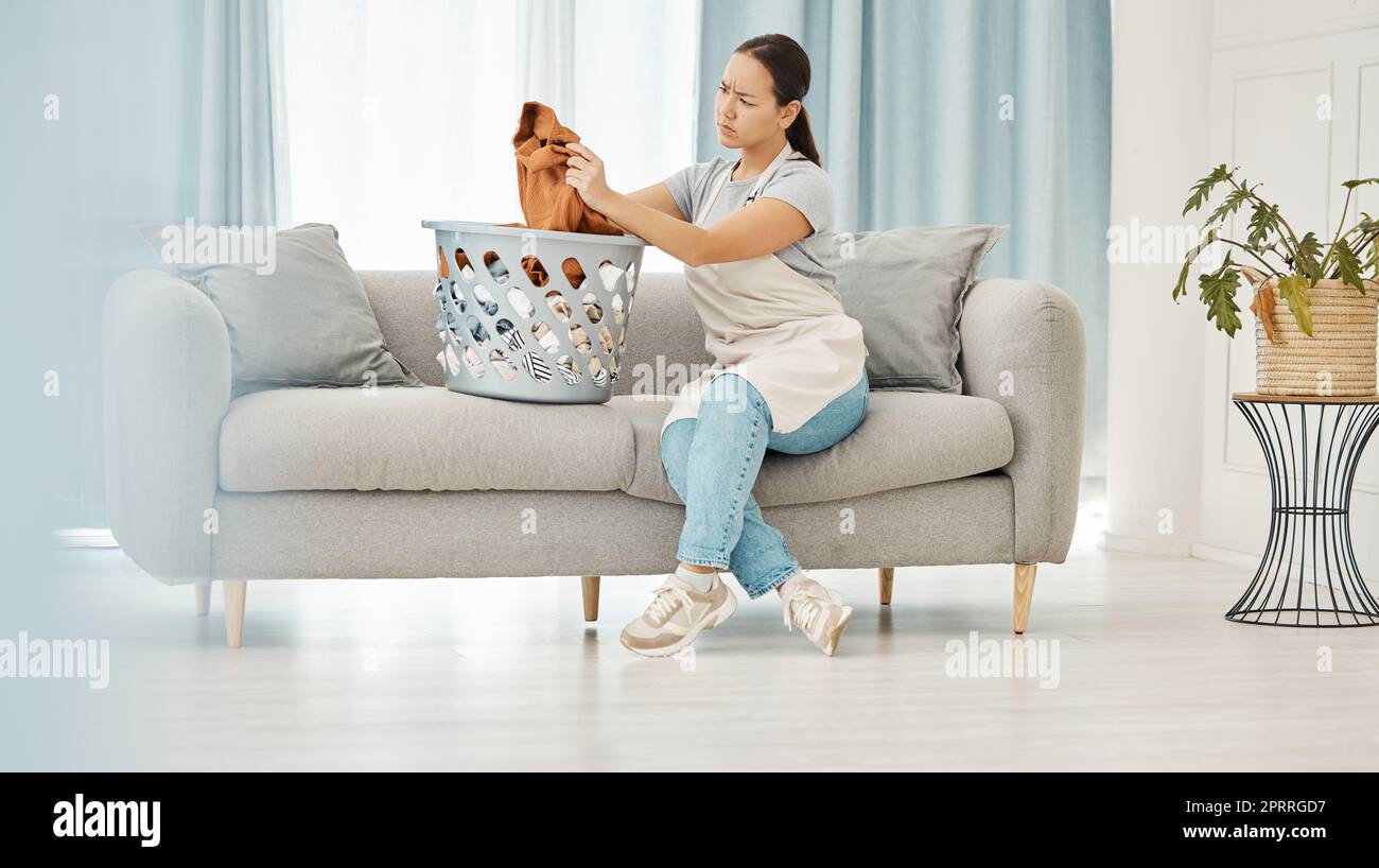 Asian woman, laundry or clothes basket on sofa in house living room and looking confused at dirty stains. Stress, anxiety or worry of person with washing clothing or cleaning fabric in home interior Stock Photo