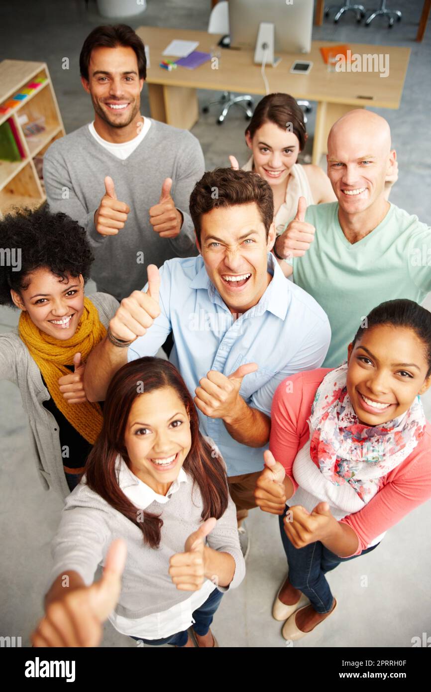 People who are passionate. High angle view portrait of a group of multi-ethnic people holding thumbs up in a work environment. Stock Photo