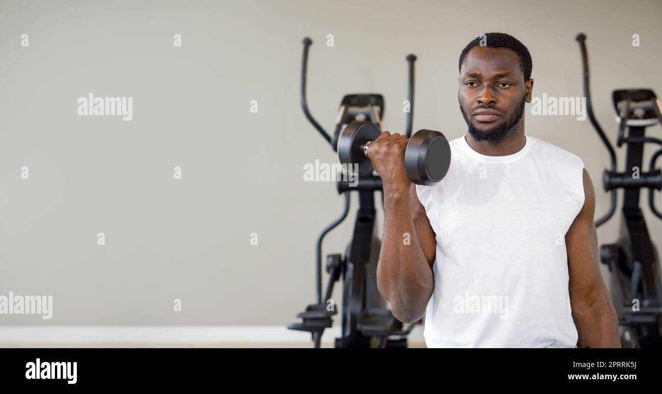 Young short curly black hair man with moustache and beard working on his biceps, lifting barbell with one hand. There are cardio machines in the gym. Stock Photo
