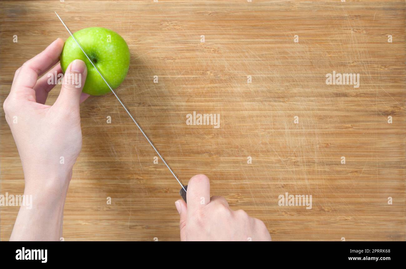 Closeup hand holding knife cutting green apple on a wooden chop board. Top View. Stock Photo