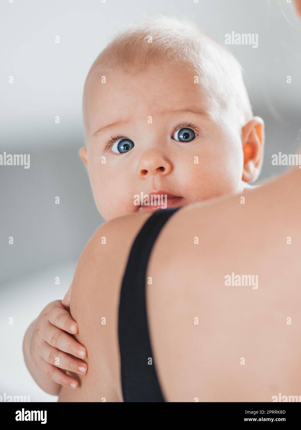Portrait of sweet baby resting in mothers arms, looking at camera, touching mama shoulder. New mom holding little kid, embracing child with tenderness, love, care. Motherhood concept Stock Photo