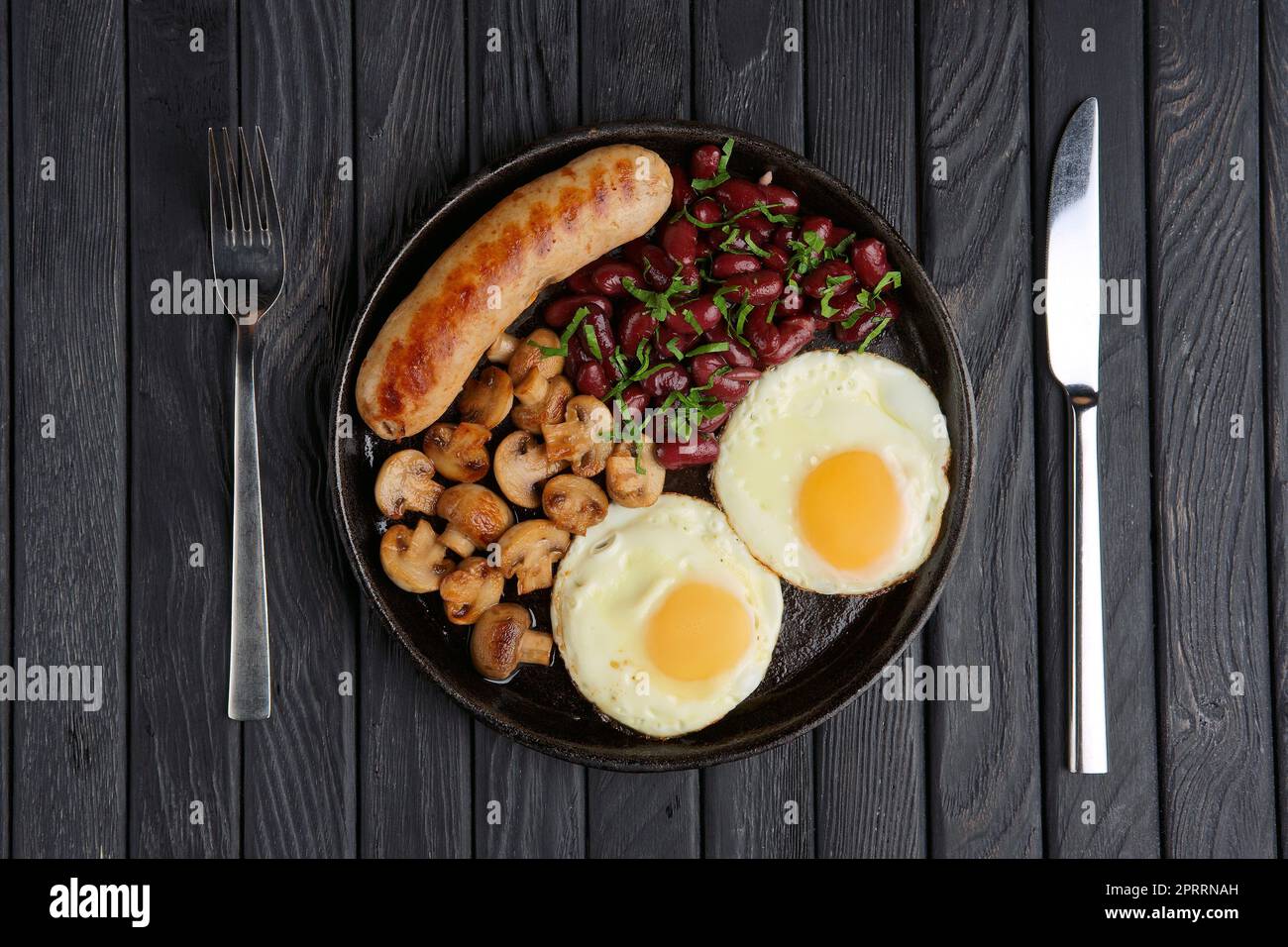 Castiron frying pan with fried eggs, sausage, mushroom and beans on wooden table. Top view