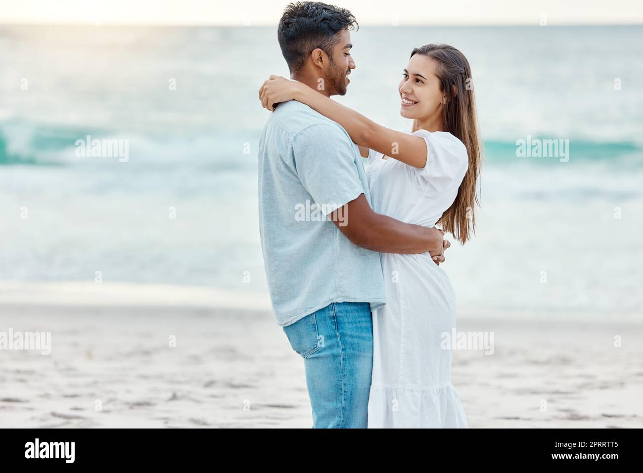 Couple love at beach, look in eyes and hug, with sunset over ocean in nature background or scene. Happy young, man and woman on sea sand smile together, sun setting over waves in backdrop or horizon. Stock Photo