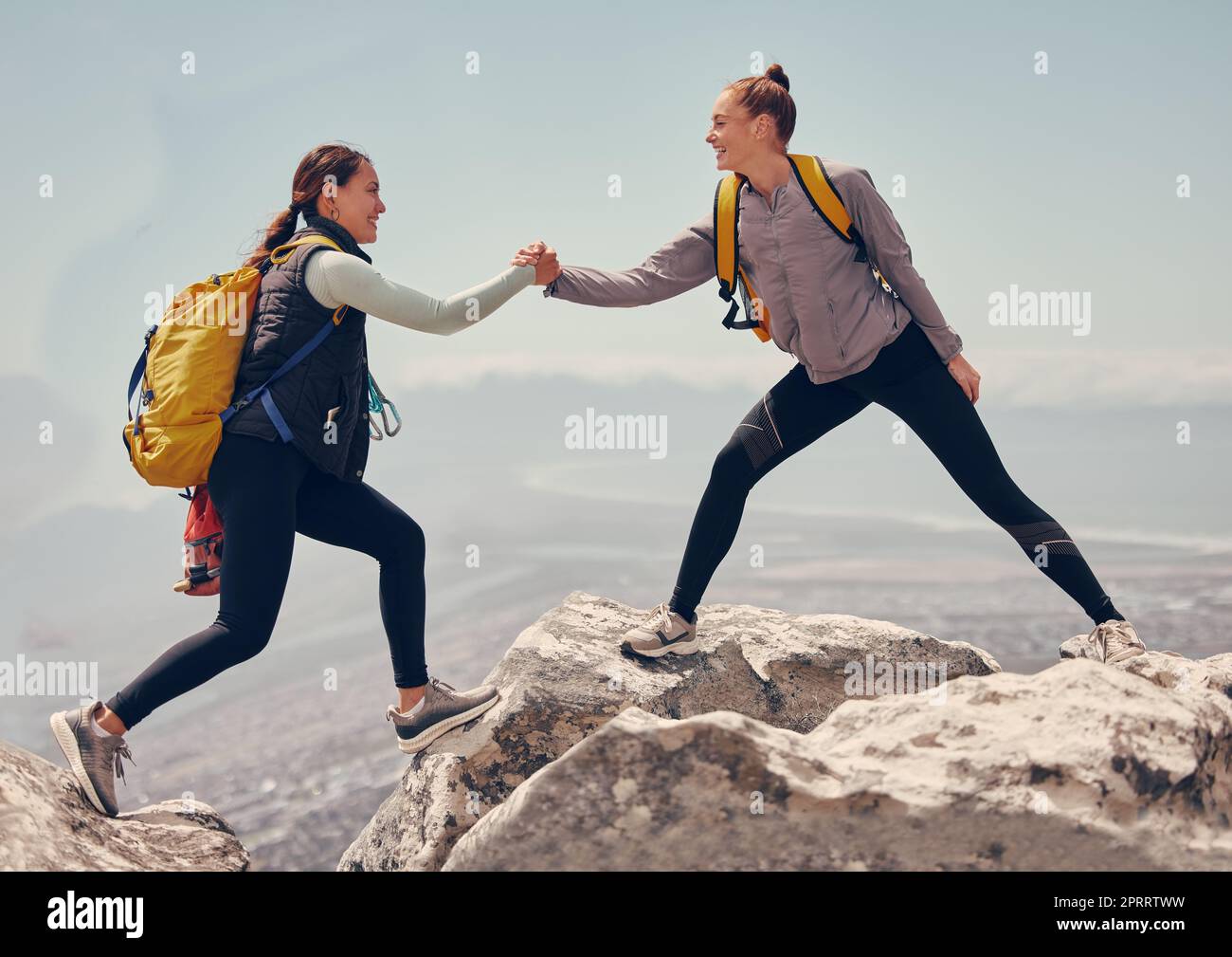 Happy women help while hiking up a rocky mountain in nature with backpack. Females friends exercise in nature park climbing and jumping while with sportswear training or trekking together outdoors Stock Photo