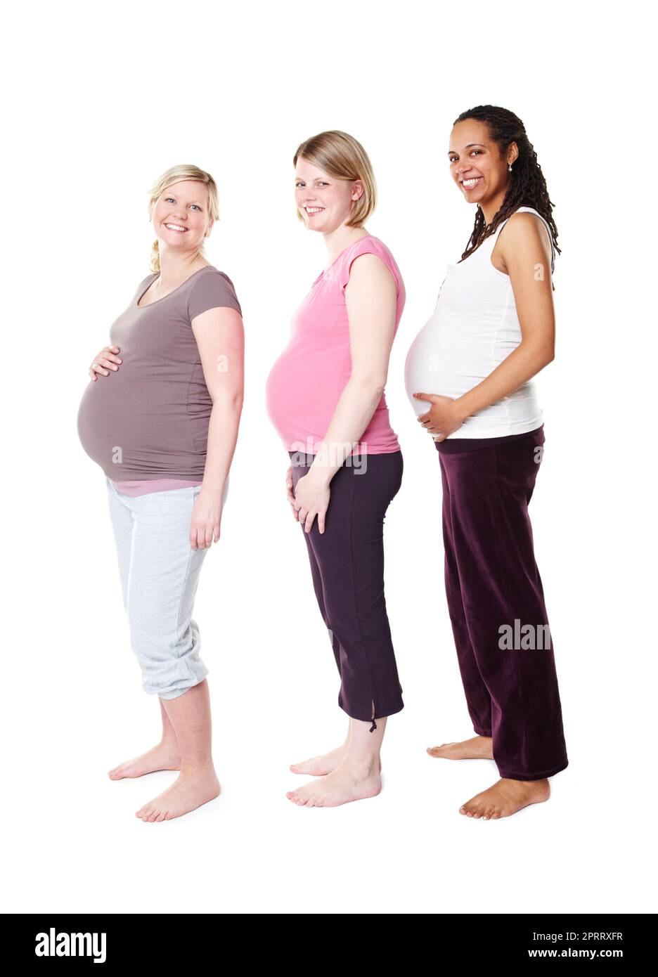 Comparing their baby bumps. Three pregnant women smiling broadly at the camera while standing against a white background holding their stomachs. Stock Photo