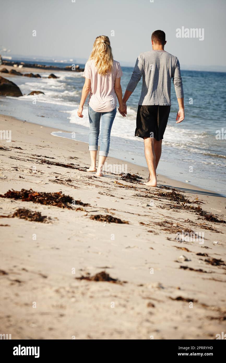 The perfect place for a walk. Rearview shot of a young couple walking hand in hand along a sandy beach. Stock Photo