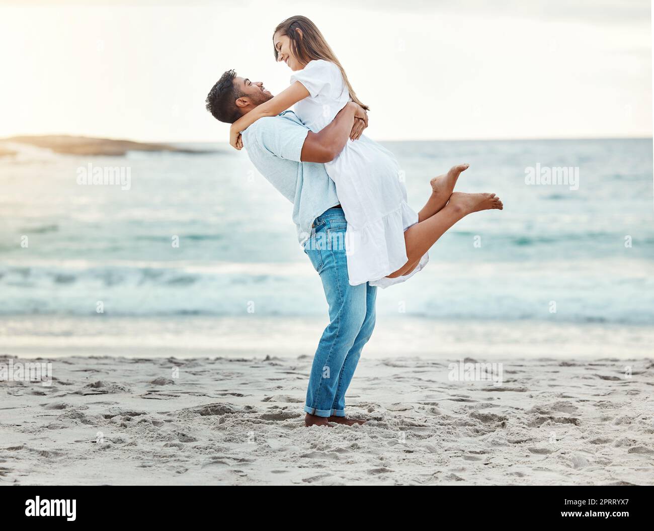Man at beach lift woman with love, smile with sun setting over the horizon or background. Young couple travel to ocean on vacation, happy together with sunset over sea or waves in nature backdrop. Stock Photo