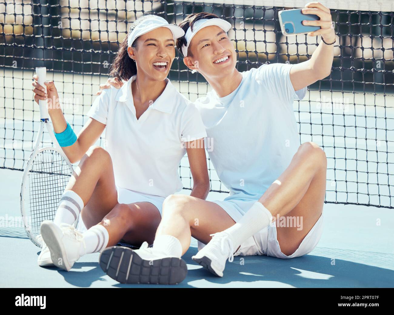 Selfie, sports or couple sitting on a tennis court resting after exercise, practice or training. Wellness, fitness or health with an interracial man and woman with sport racket taking a phone picture Stock Photo