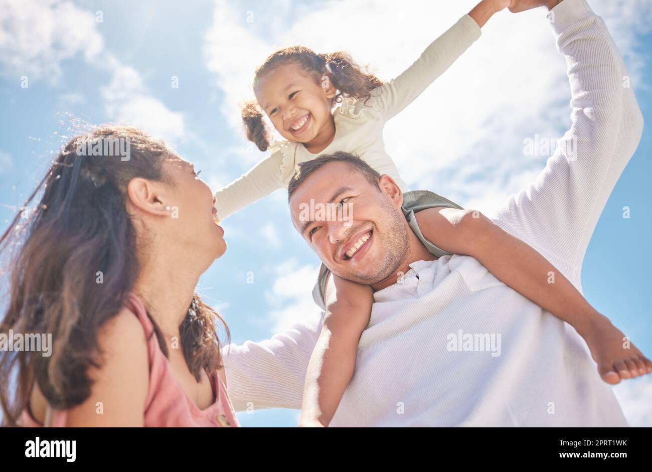 Children, family and love with a girl on the shoulders of her dad outside with her mother watching on against a blue sky. Kids, happy and smile with a daughter having fun with her parents outdoor Stock Photo