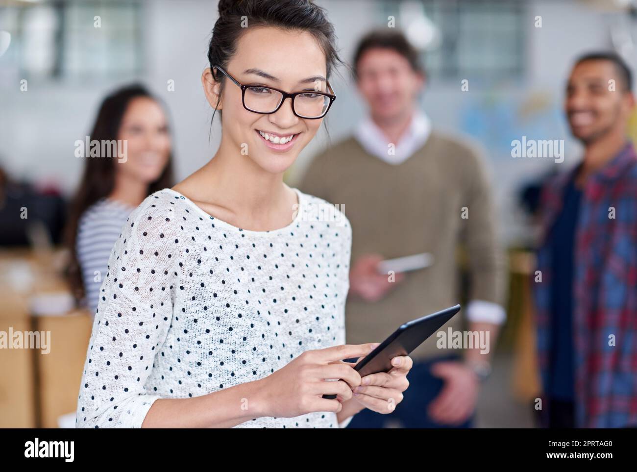 Our business is tech savvy. Friendly young woman holding a digital tablet with smiling staff behind her. Stock Photo