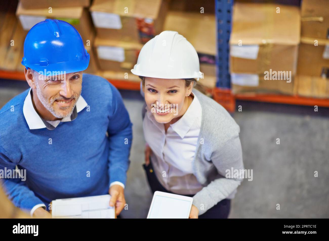 We have what it takes. Overhead shot of two people wearing hardhats smiling at the camera in a warehouse. Stock Photo
