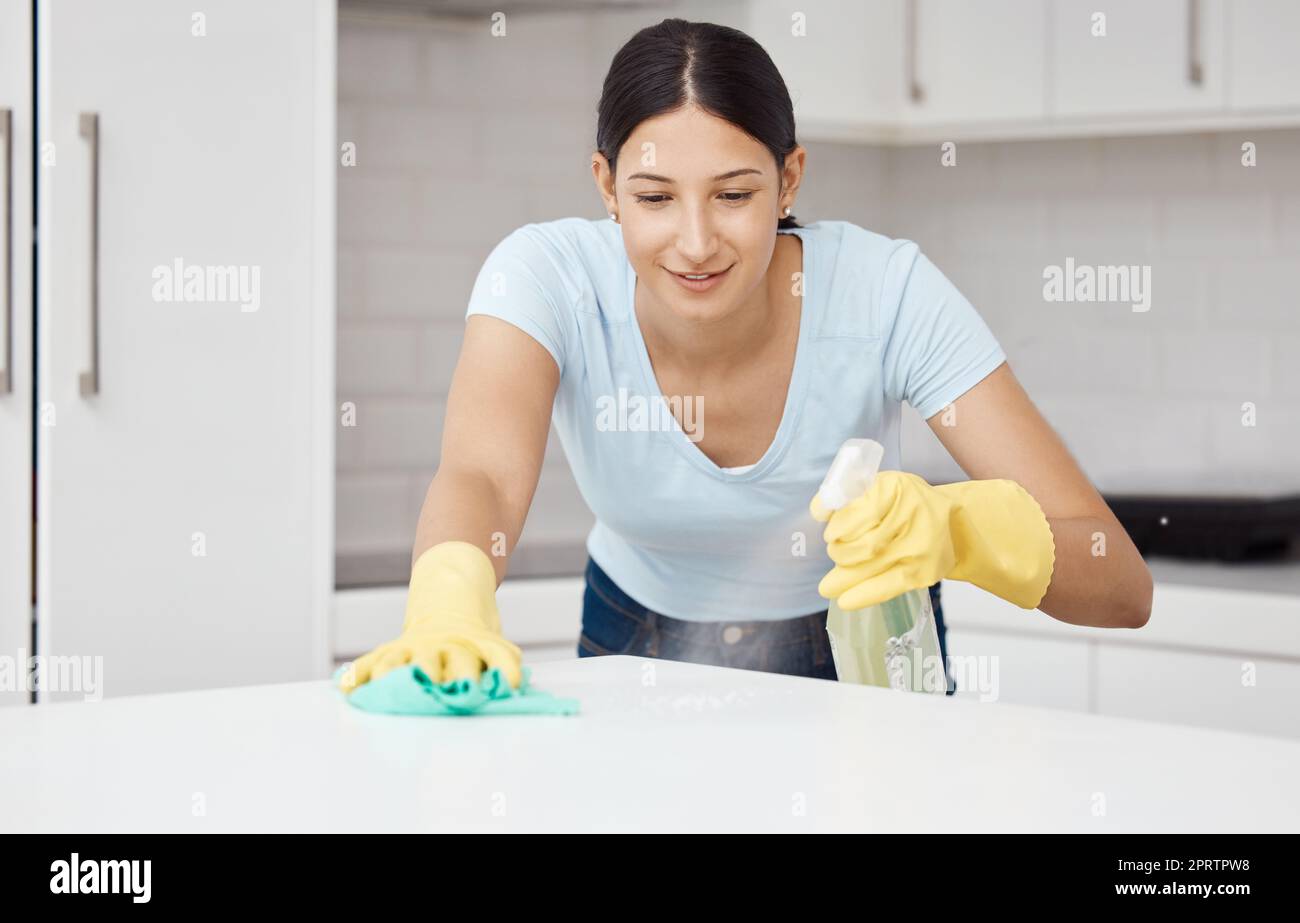 Cleaning service, table and cleaner in the kitchen working with spray bottle to scrub messy dirt with a cloth and soap detergent. Happy, woman and employee on housekeeping job in gloves with products Stock Photo