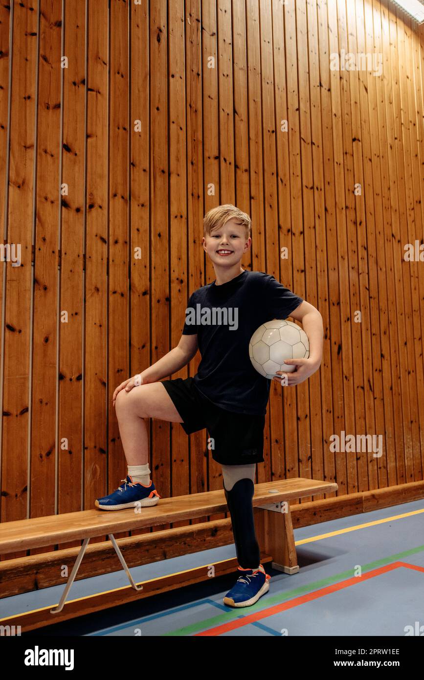 Smiling boy with amputated leg holding soccer ball while standing by wooden wall at sports court Stock Photo