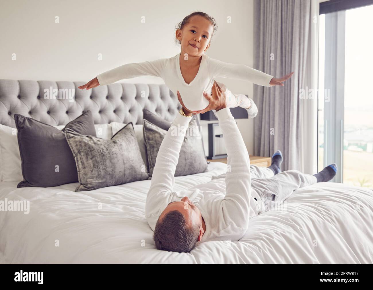 Father lifting child in air on bed, playing and bonding in bedroom of house. Love portrait, care and trust of male parent holding happy kid up in house spending free time together having fun at home. Stock Photo