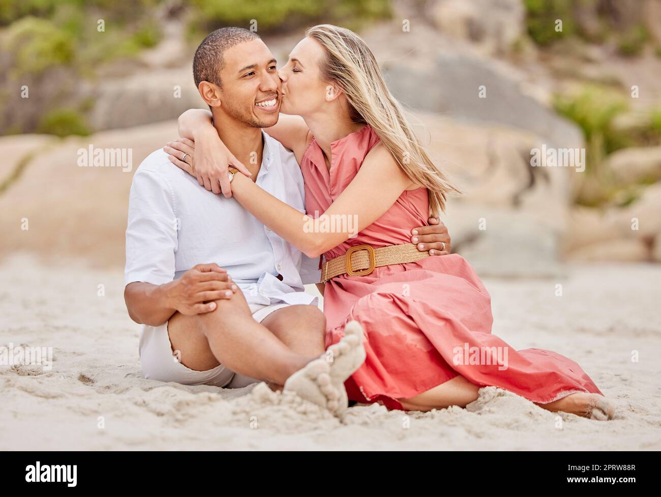 Kiss, love and couple on beach sand with happy smile for holiday or vacation together. Healthy happiness and interracial man and woman or people relax on ground for wellness lifestyle Stock Photo