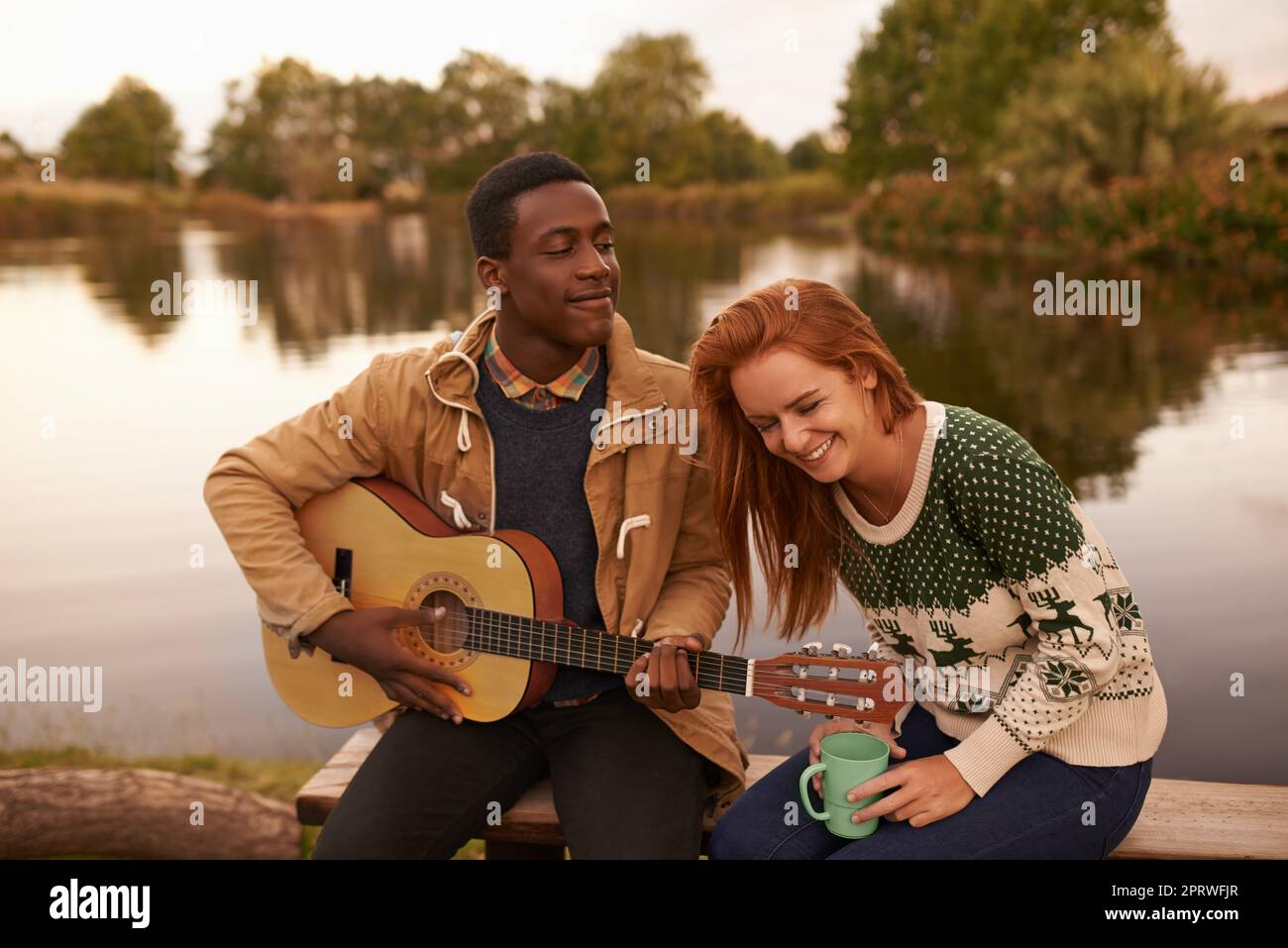 He knows that the key to her heart is laughter. a teenage boy serenading a beautiful girl while sitting beside a lake. Stock Photo