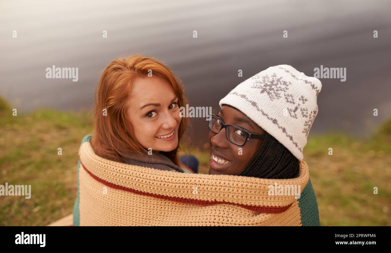 A cosy catch up by the lake. High angle portrait of two happy teenage girls huddled under a blanket beside a lake. Stock Photo