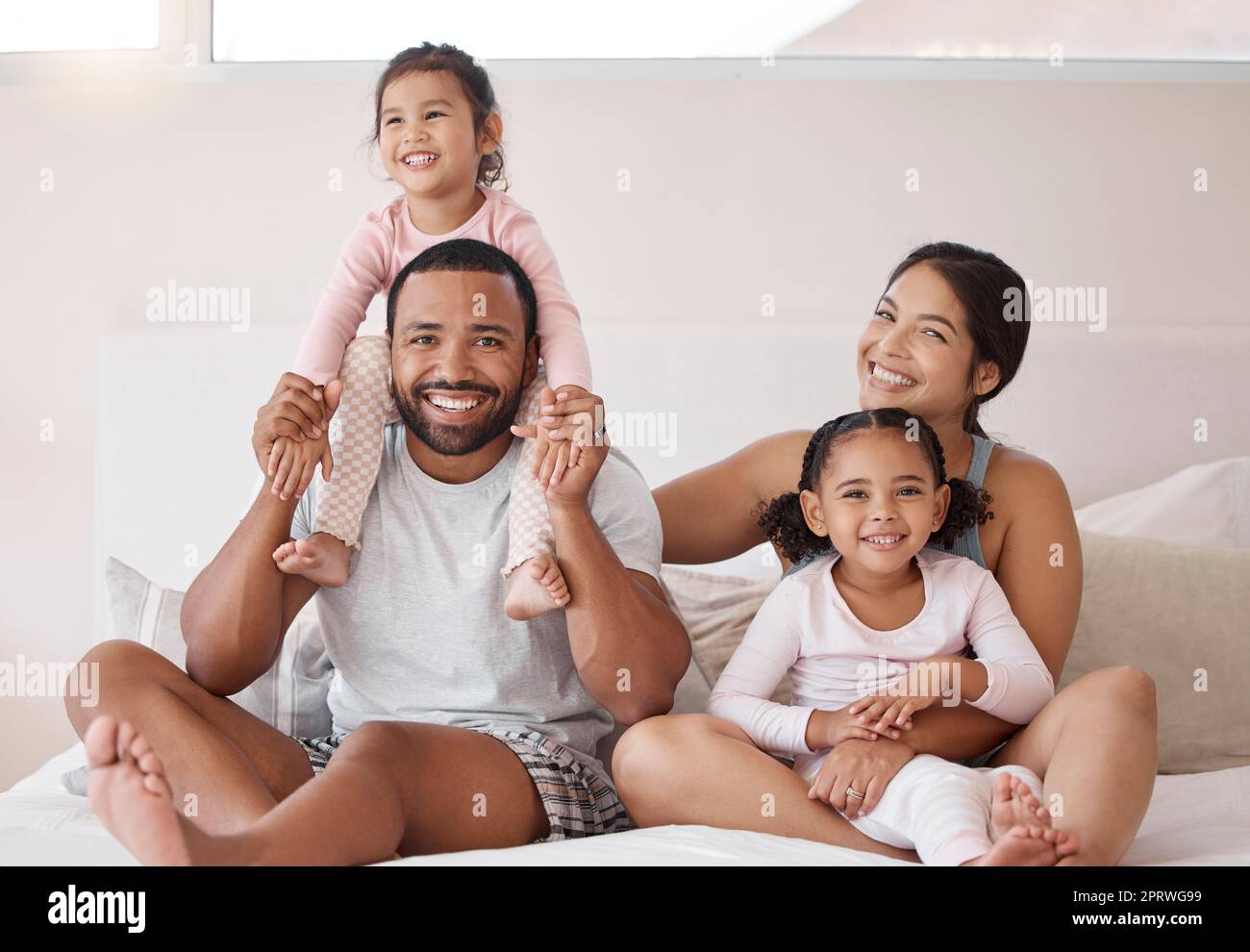 Family, children and love with a man and woman sitting on a bed with their kids at home. Happy, smile and girl with her sister, mother and father relaxing together in a bedroom in their house Stock Photo