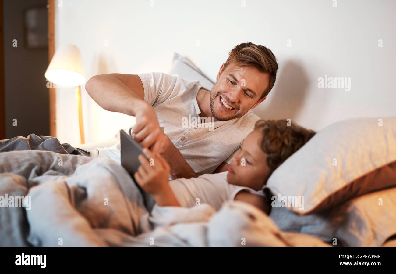 I know the answer to this one. a young boy using a digital tablet while lying in bed with his dad. Stock Photo