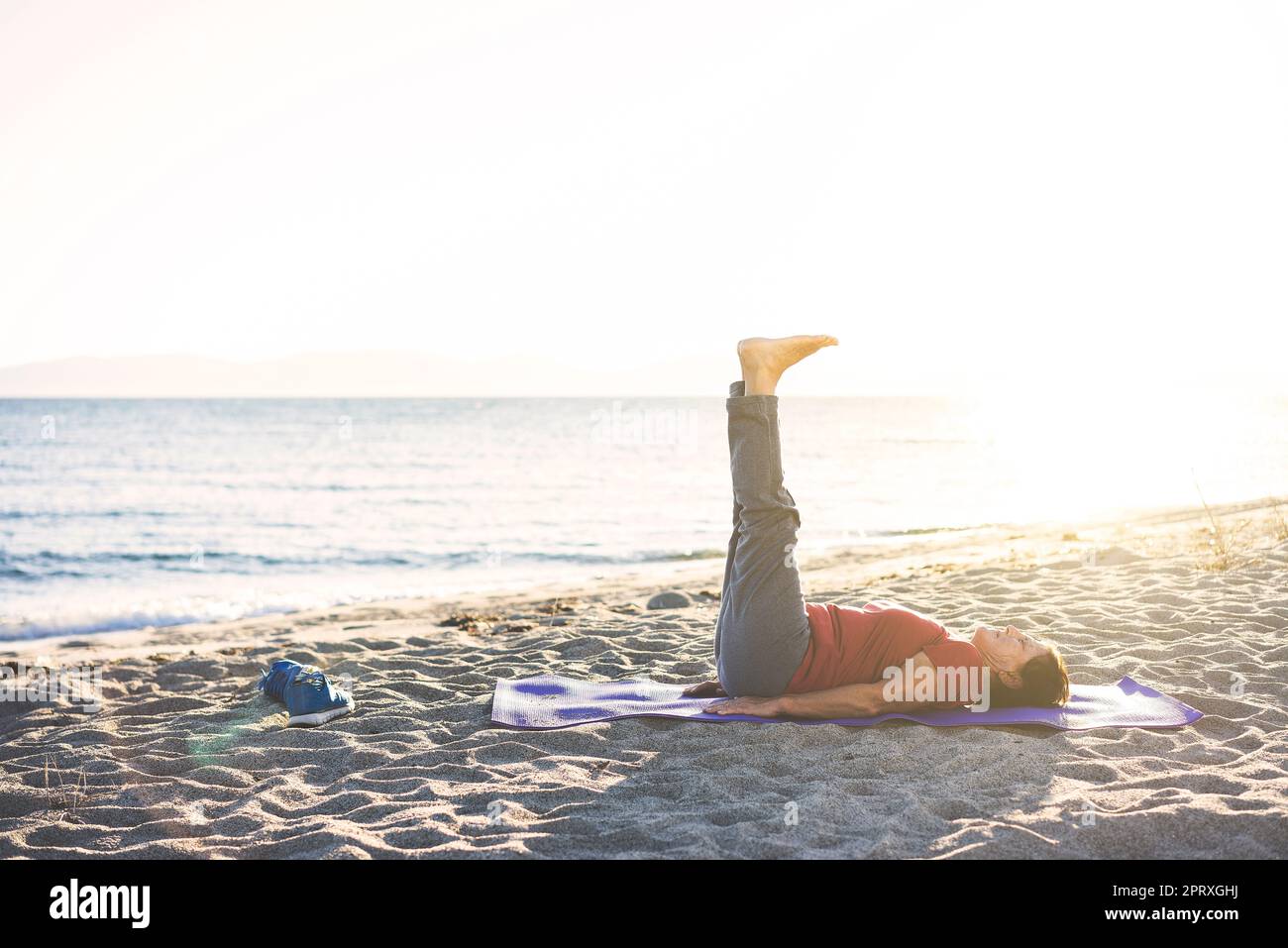Senior woman at the beach laying on yoga mat, exercising. Doing leg raises. Stock Photo