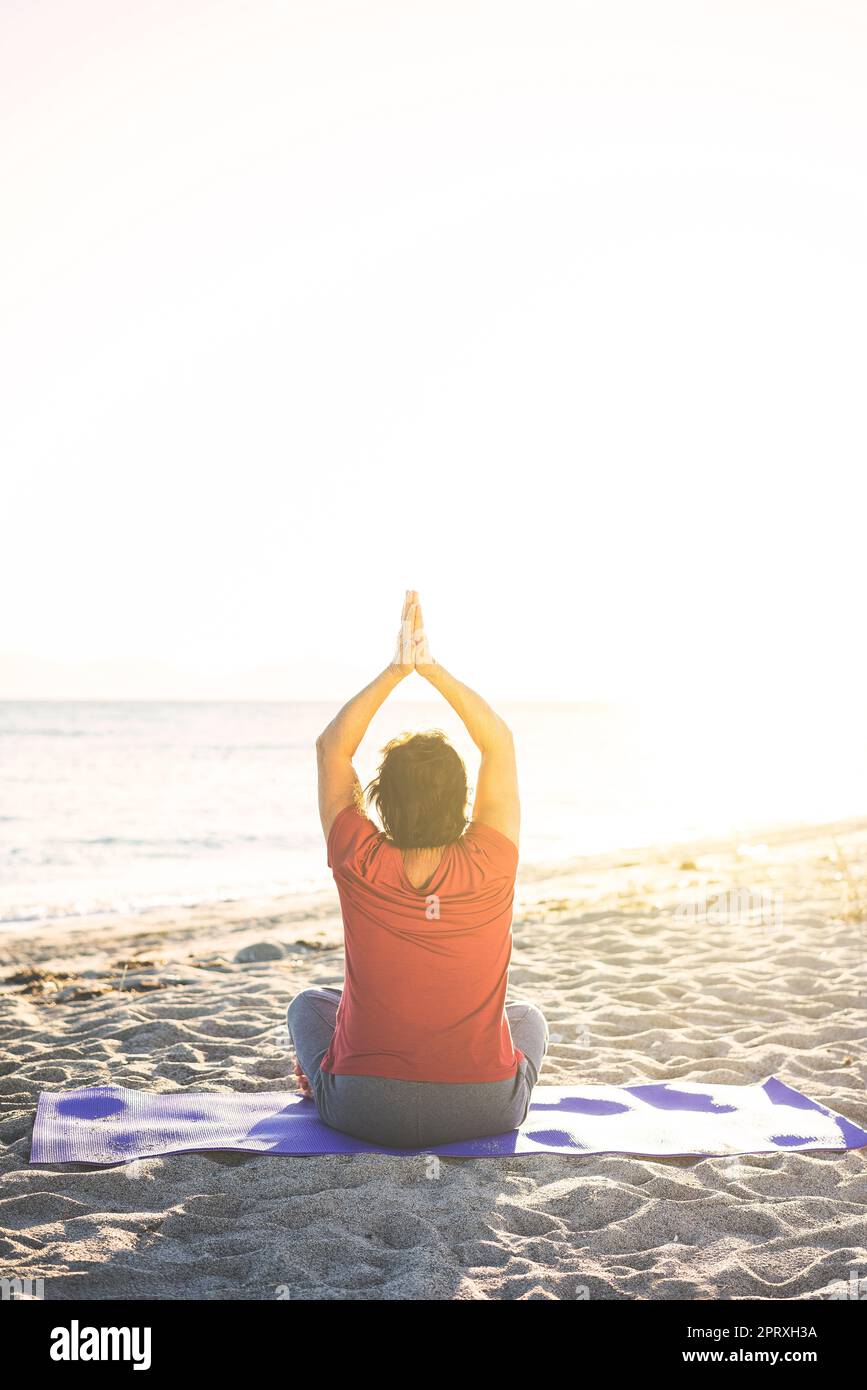 Senior woman at the beach, doing exercise on yoga mat, meditating. Shot from back. Stock Photo