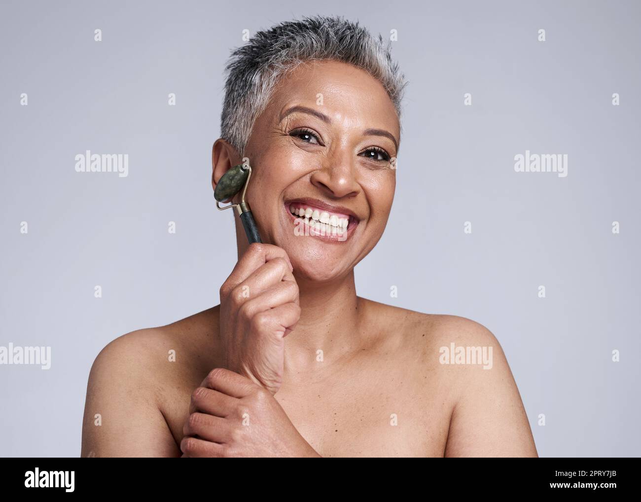 Woman, roller and face for skincare beauty, health or exfoliate for cosmetic wellness by wall. Happy, senior model and crystal for dermatology massage Stock Photo