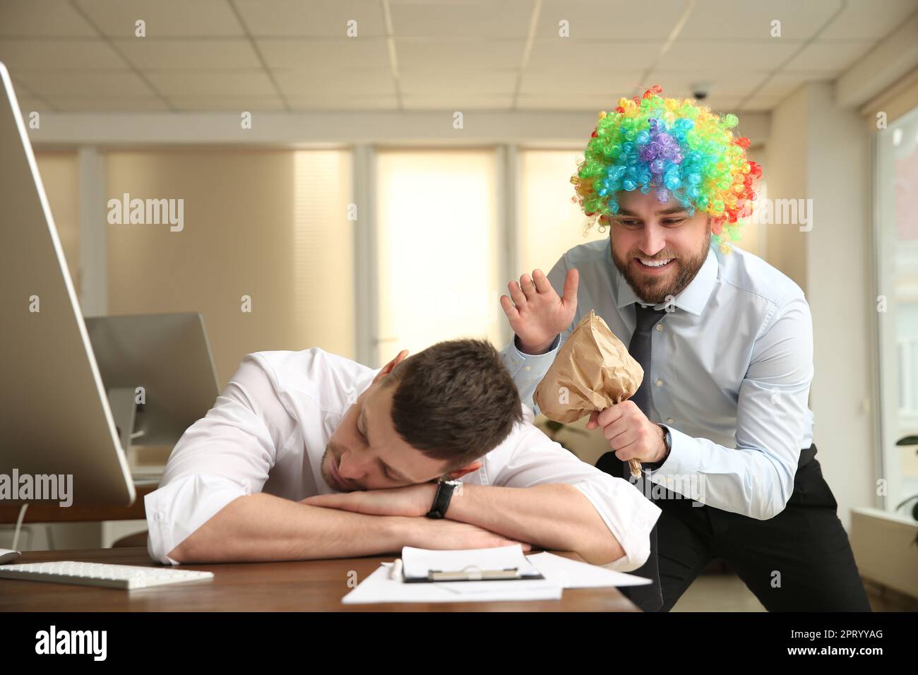 Young man popping paper bag behind his sleeping colleague in office