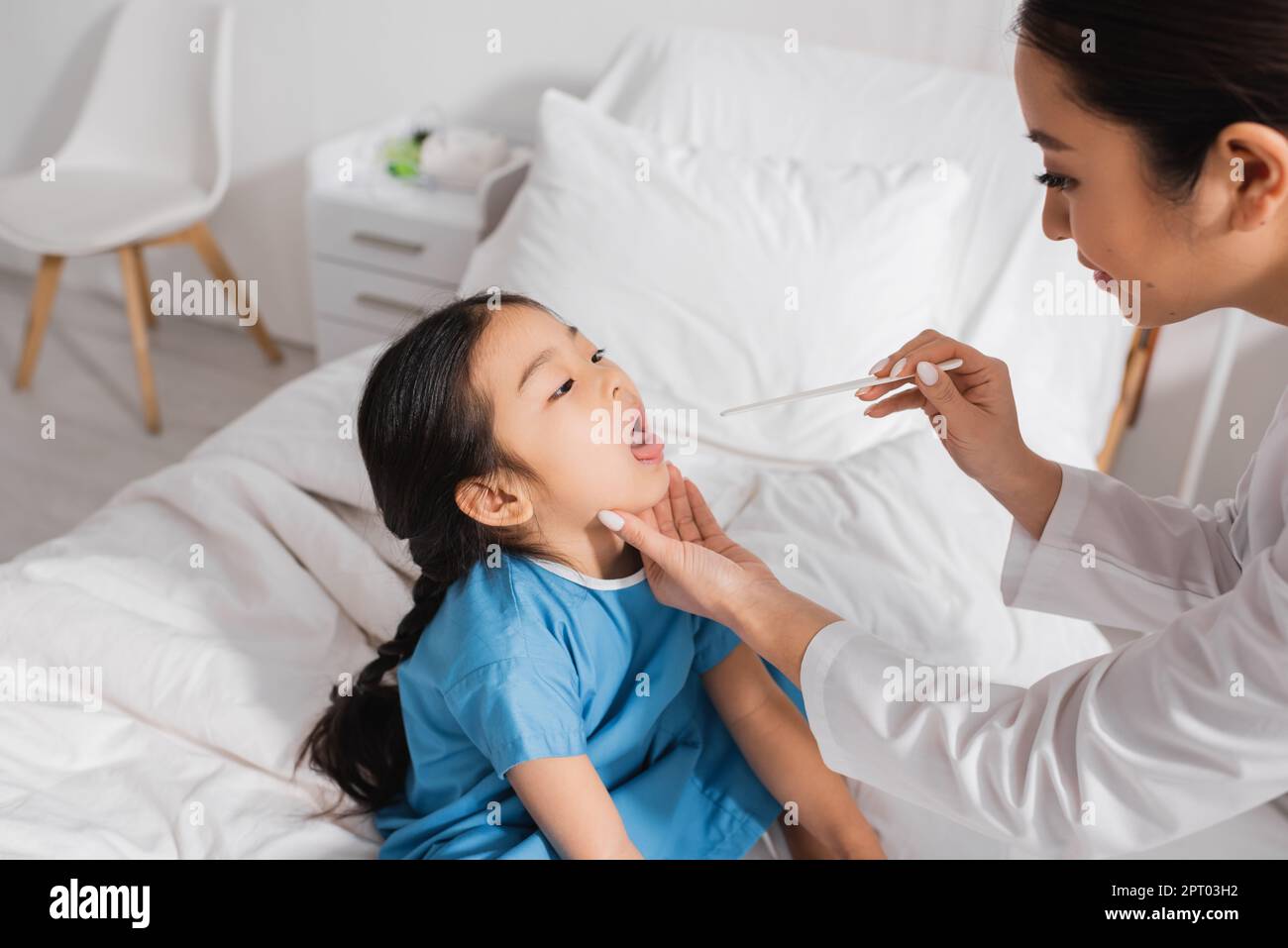 asian girl sitting with open mouth near pediatrician with tongue depressor in hospital ward