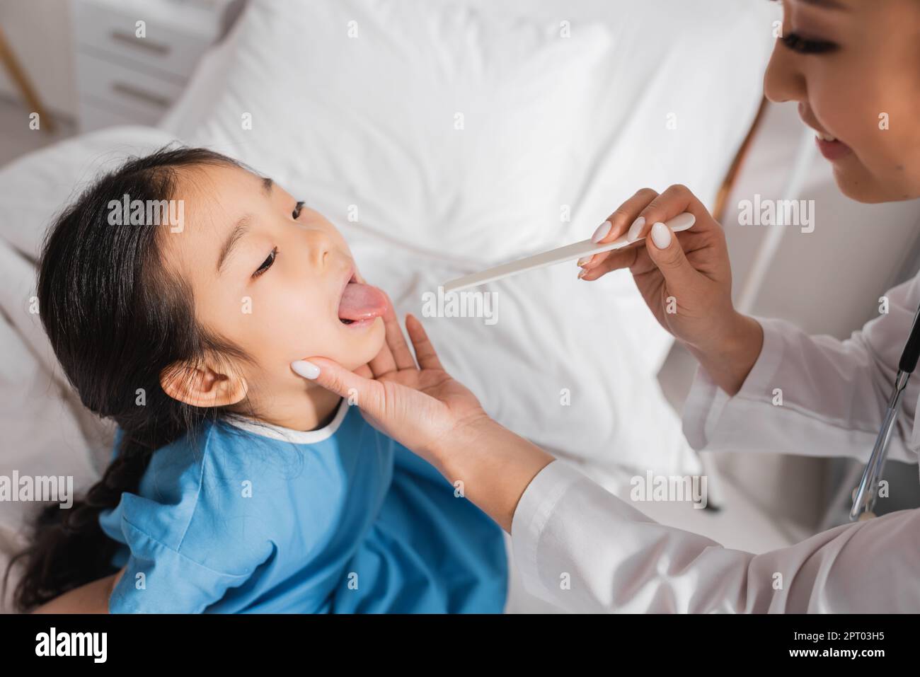 asian girl opening mouth near smiling doctor with tongue depressor in
