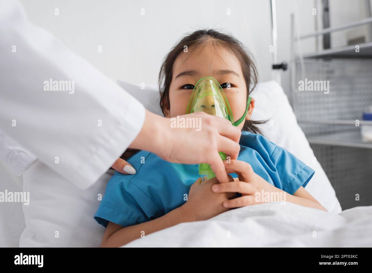 little asian girl breathing in oxygen mask near doctor in hospital ward