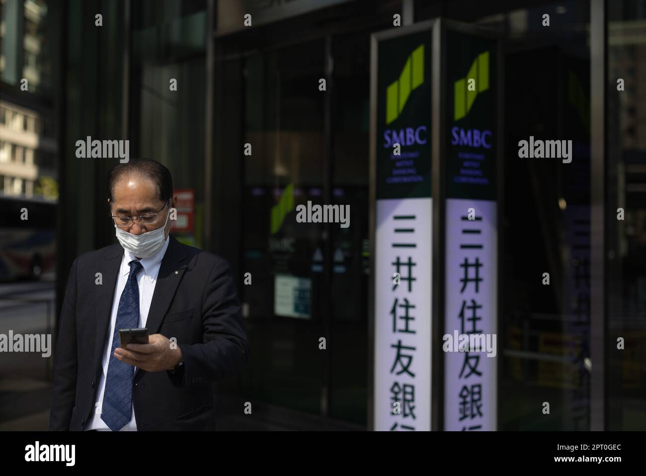 Japanese businessman with a face mask looks at his mobile phone in front of Sumitomo Mitsui Bank in central Tokyo. Stock Photo