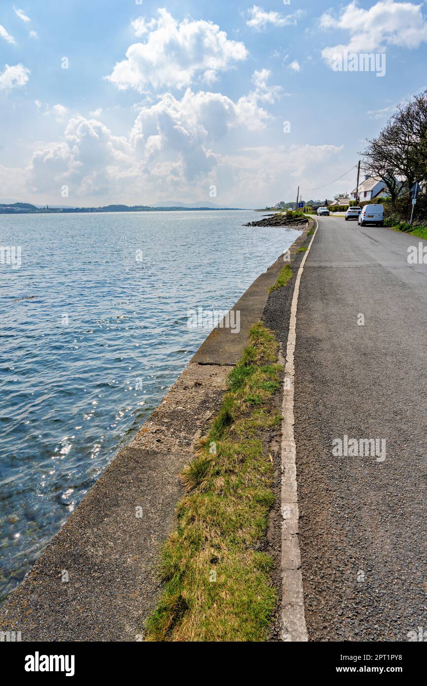 A tranquil and scenic view of the Menai Straits near Anglesey, Wales with blue sky, white clouds, green land and trees beside a winding coastal road. Stock Photo
