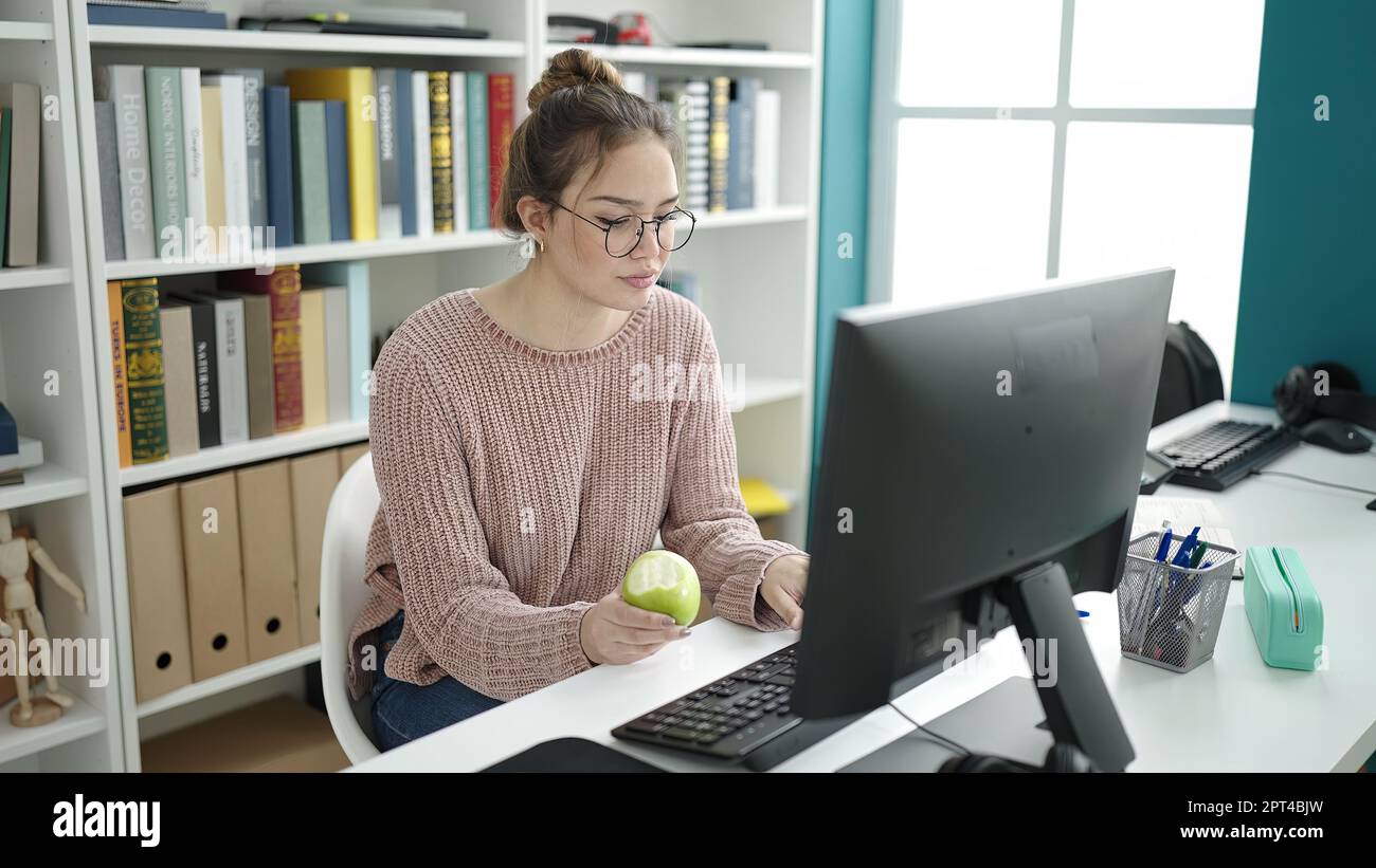 Young beautiful hispanic woman student using computer eating apple at ...