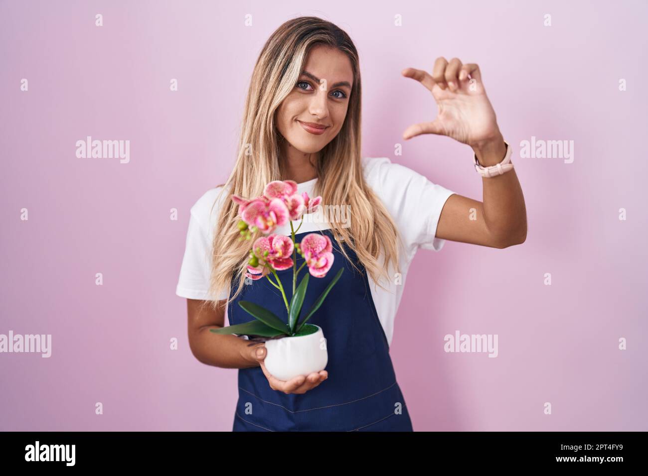 Young blonde woman wearing gardener apron holding plant smiling and confident gesturing with hand doing small size sign with fingers looking and the c Stock Photo