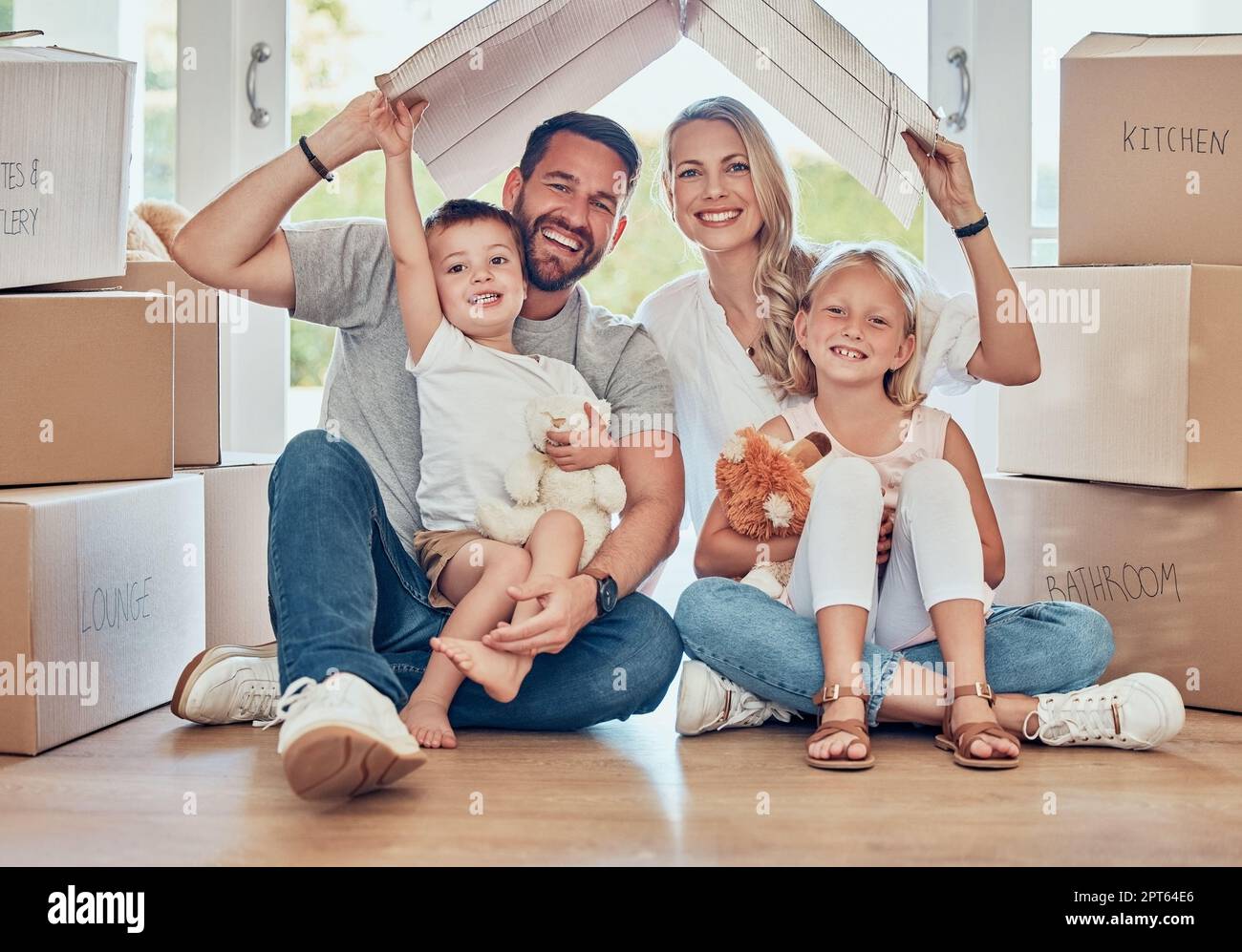 Smiling couple with little kids sitting and mockup roof over children. Cheerful family sitting together on floor in new house on relocation day. Paren Stock Photo