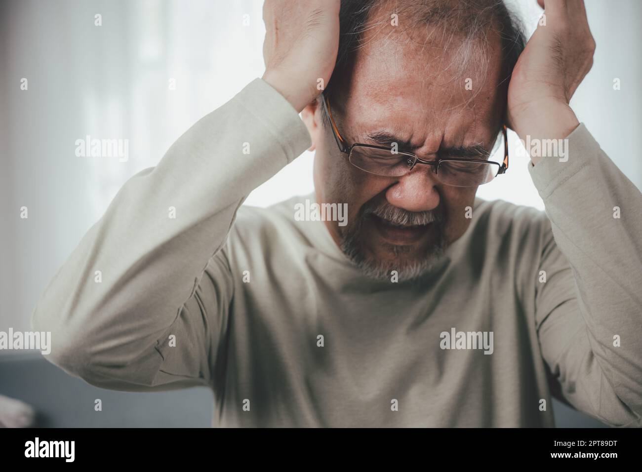 Headache. Close up of elderly holds head with hand suffering from migraine headache, Sad Asian senior man sitting on sofa feeling hurt and lonely at h Stock Photo