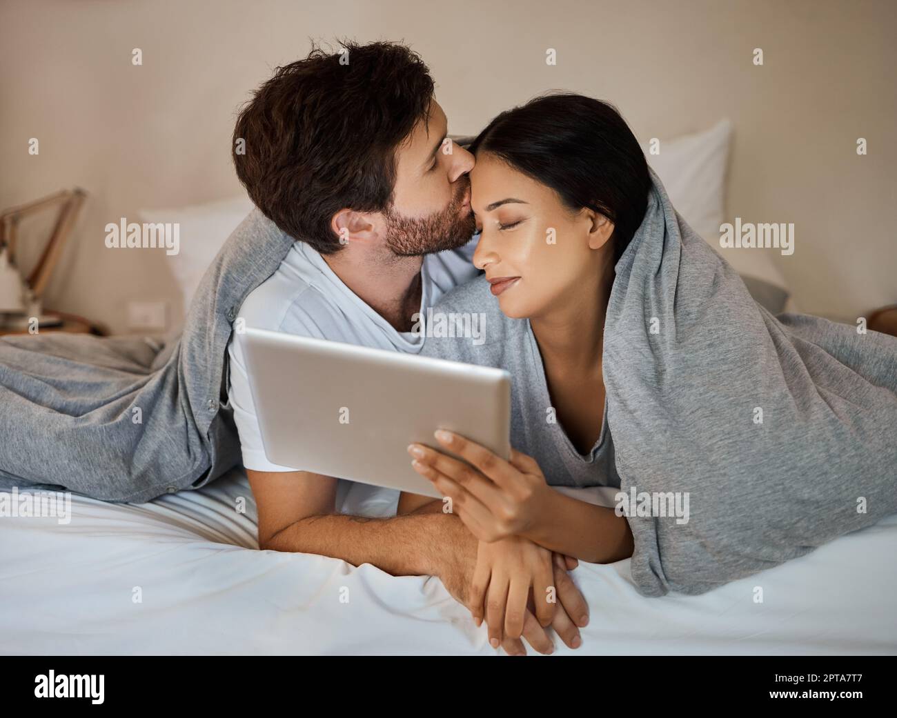 Couple, love and tablet for entertainment while lying in bed with a kiss on forehead while watching a movie, internet browsing and streaming service Stock Photo