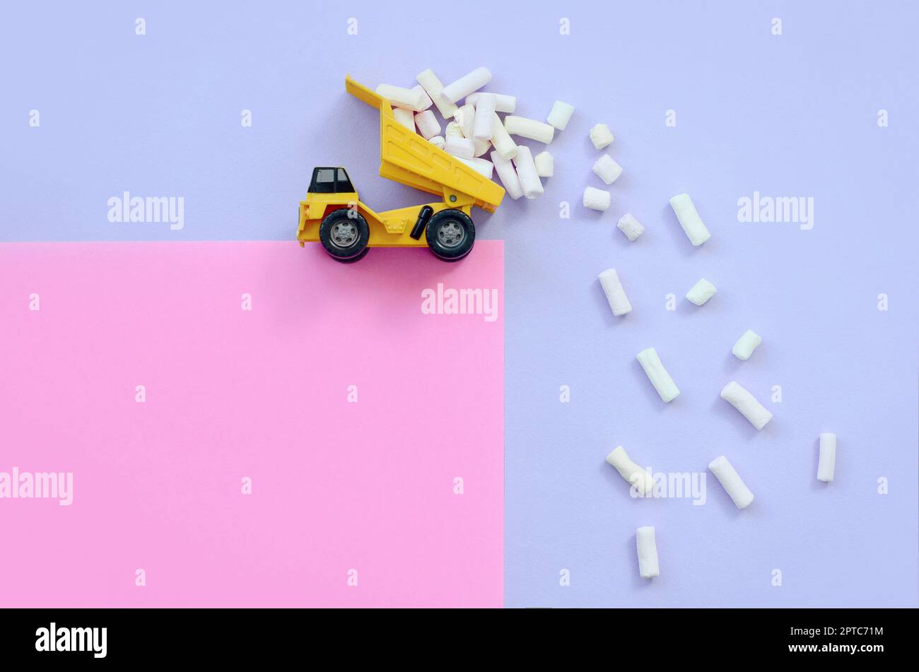 Yellow little toy dump truck throws marshmallow pieces from its raised back on a pastel violet and pink background. Flat lay minimal top view. Stock Photo