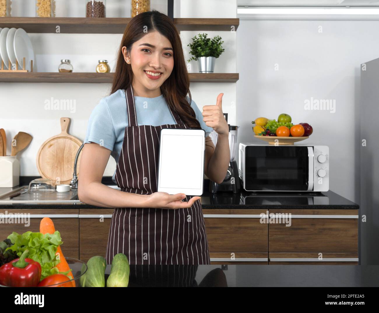 The housewife dressed in an apron  showing white screen tablet computer while raise finger thumbs up. Morning atmosphere in a modern kitchen. Stock Photo
