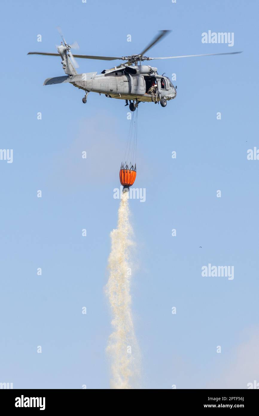 A U.S. Navy SH-60 Sea Hawk helicopter drops water from a bambi bucket over a target area during the annual Cory Iverson Wildland Firefighting exercise at the Las Pulgas Lake on Marine Corps Base Camp Pendleton, California, April 19, 2023. The Cory Iverson Wildland Firefighting exercise is an annual event that focuses on interagency cooperation between the California Department of Forestry and Fire Protection, San Diego County Sheriff's Department, and the Department of Defense. During the exercise, each entity works together to create an effective, efficient, and well trained team to respond i Stock Photo