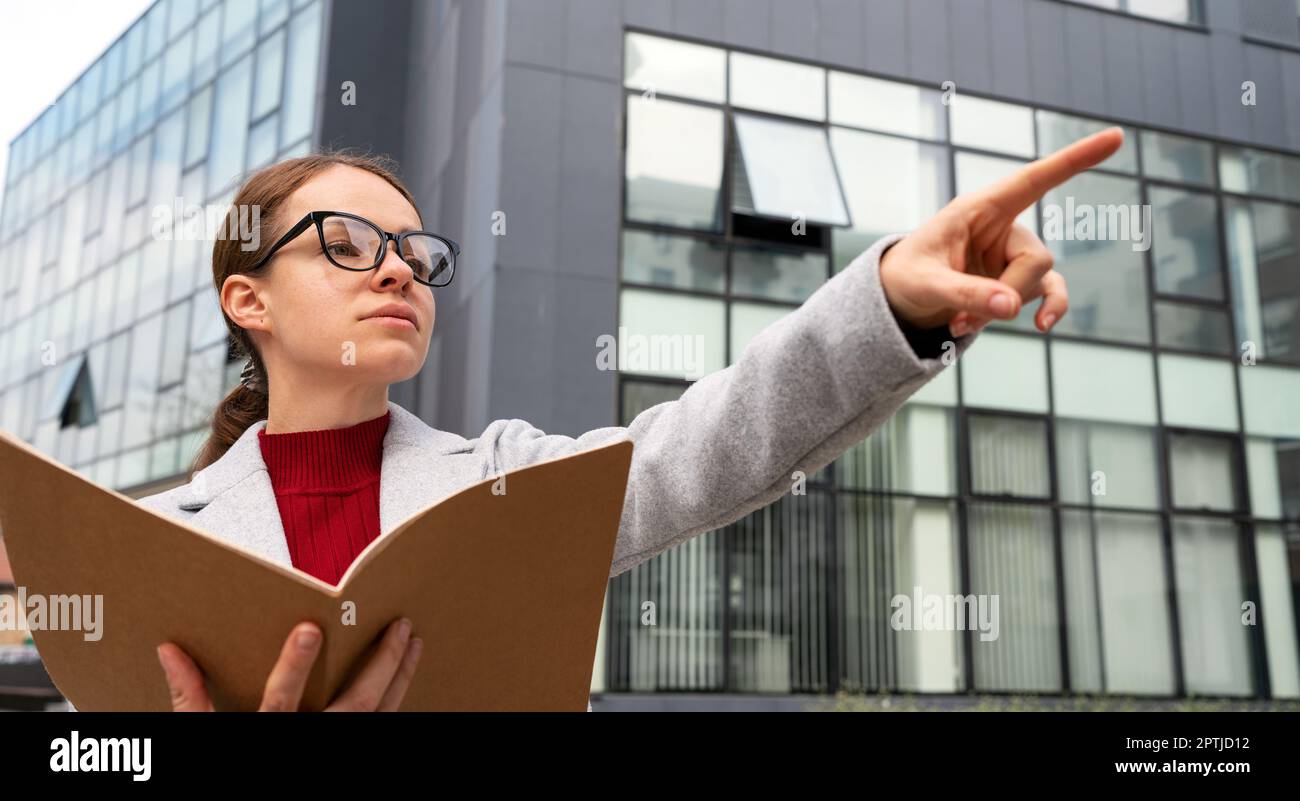 Business person woman wearing glasses holds folder with documentation and pointing hand at facility, inspecting facilities. Stock Photo