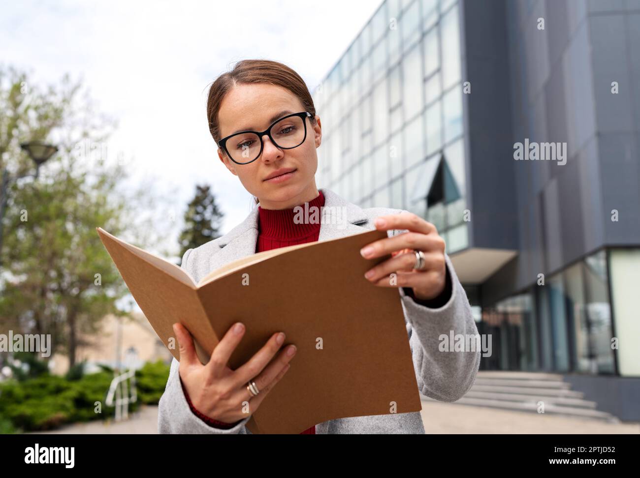 Executive manager woman wearing glasses reading project documentation outdoors standing near near office building. Stock Photo
