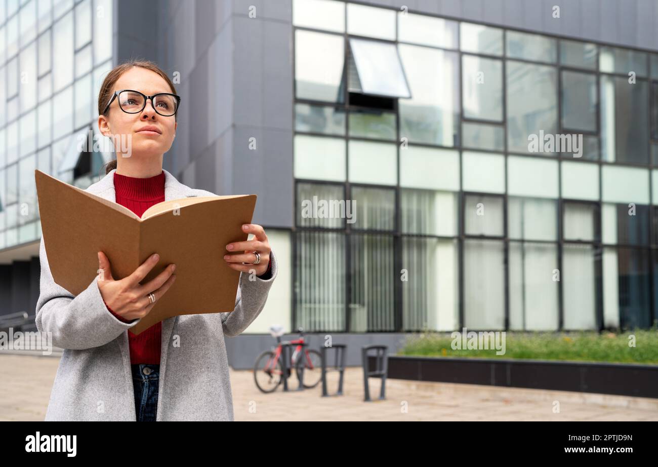 Executive manager woman wearing glasses holding folder with documentation in her hands, standing outdoors next to office building. Stock Photo